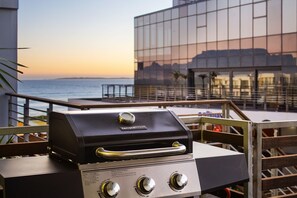 A terrace with a grill, overlooking a body of water and a modern building. - Eden On The Bay 118 (Cape Town)