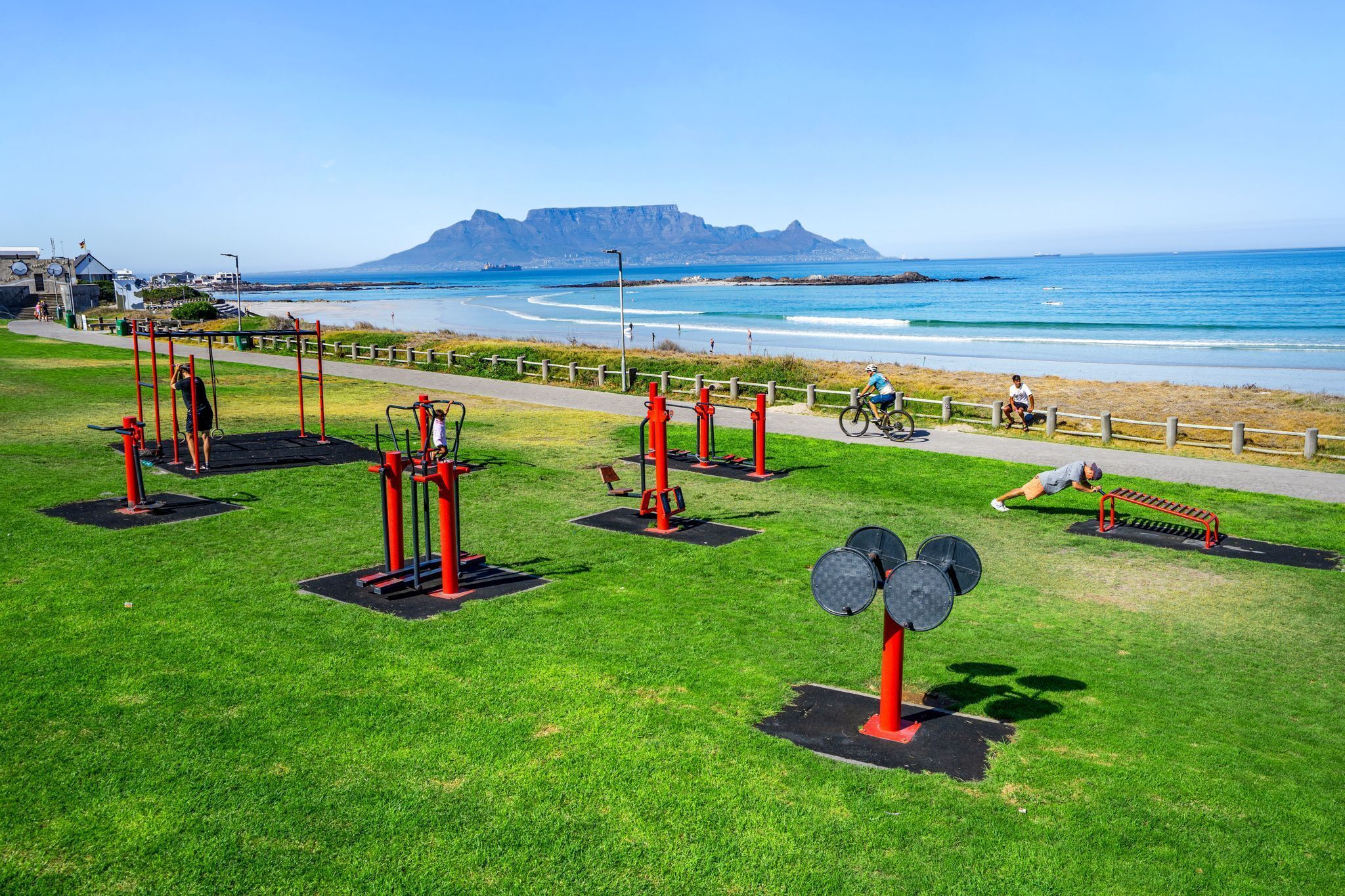 Un gimnasio al aire libre con equipo de ejercicio, un sendero pavimentado y vistas al océano y una montaña al fondo.