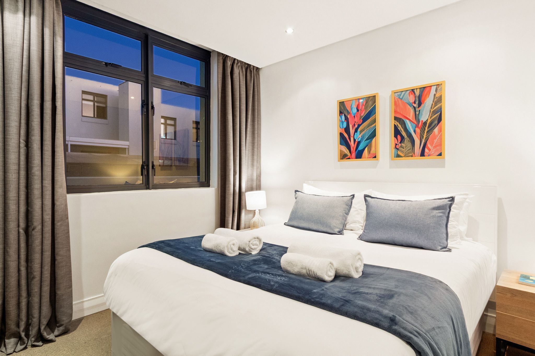 Habitación de hotel con una cama grande, dos almohadas, una colcha azul y dos toallas enrolladas. Hay dos cuadros abstractos en la pared y una ventana con cortinas.
