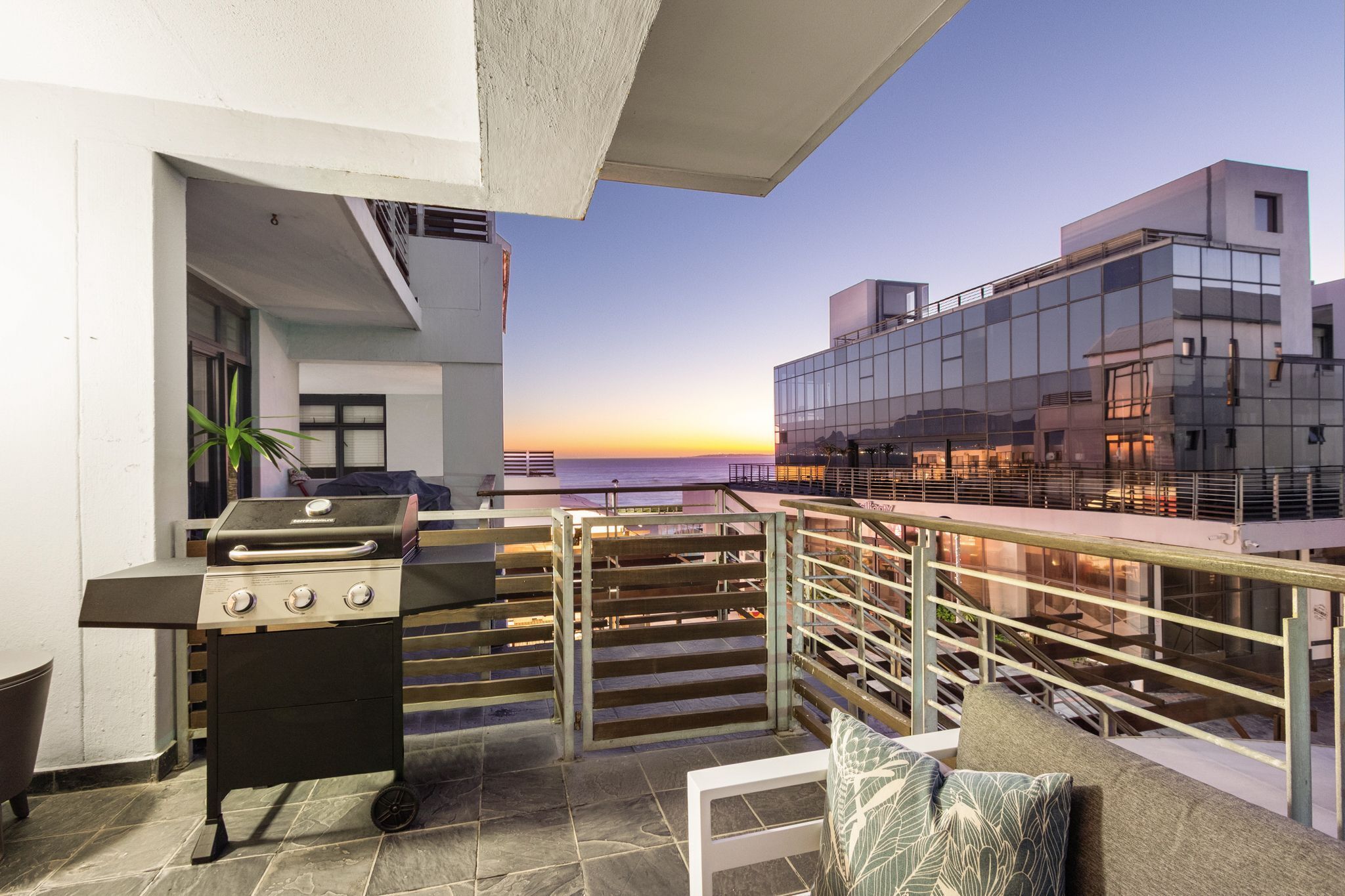 A rooftop terrace with a barbecue grill, a seating area with cushions, and a view of the sunset over the ocean.