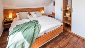 A modern bedroom with a wooden bed frame, white bedding, and a green throw blanket. There are two bedside tables with lamps, a wooden wardrobe, and a small shelf with decorative items. - Zum Brandenburger Tor 12 (Damme, Dümmer)