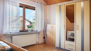A bedroom with a wooden ceiling, a window with sheer curtains, a radiator, a wardrobe with a mirror, and a bed with a plaid blanket.