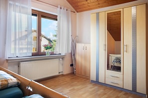 Ein Schlafzimmer mit Holzboden, einem Fenster mit transparenten Vorhängen, einem Heizkörper, einem Kleiderschrank mit Spiegel und einem Bett mit Karo-Decke.