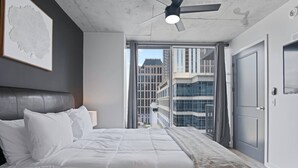 Un dormitorio moderno con una cama grande, un ventilador de techo y vistas de edificios altos a través de un ventanal.