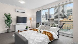 Habitación de hotel moderna con una cama grande, televisión, planta en maceta y vista a la ciudad a través de amplios ventanales.