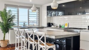 Een moderne keuken met een groot eiland, witte barkrukken, een potplant en uitzicht op zee door de ramen.