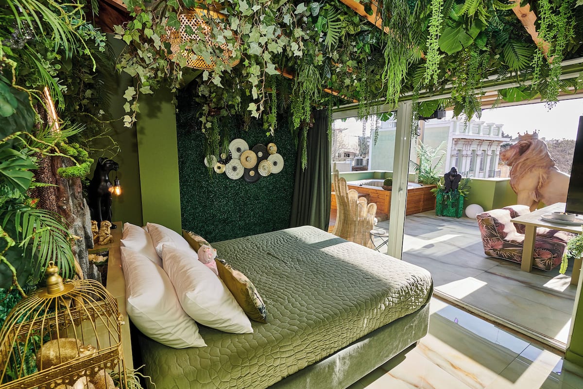 A room with a bed, green walls, hanging plants, and a large window.