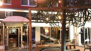 An outdoor seating area with tables and chairs, a tree, and a building with signage in the background. - Hotel Sittardsberg (Duisburg)