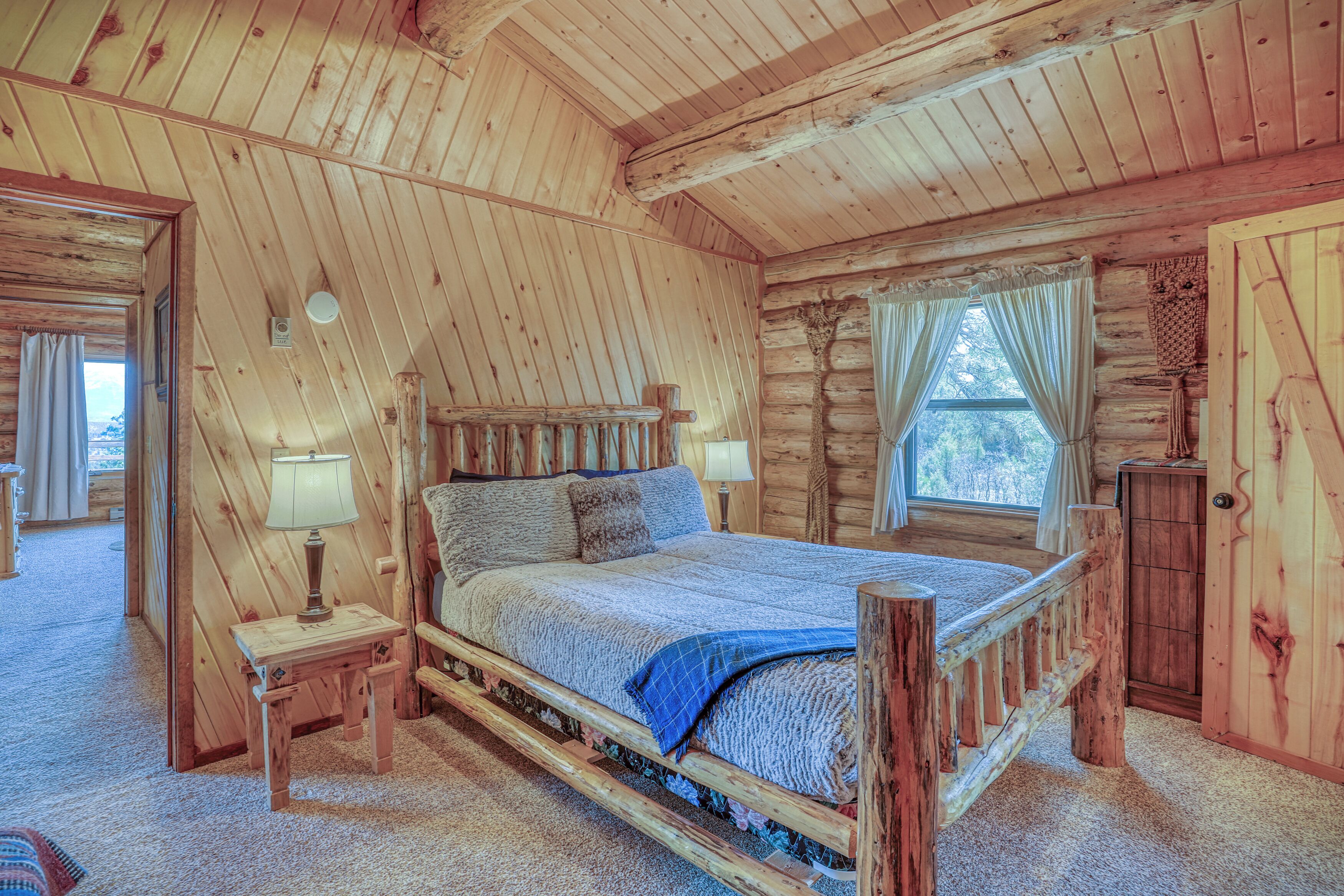 A log cabin bedroom with a wooden bed, two bedside tables with lamps, a window with curtains, and a wooden wardrobe.