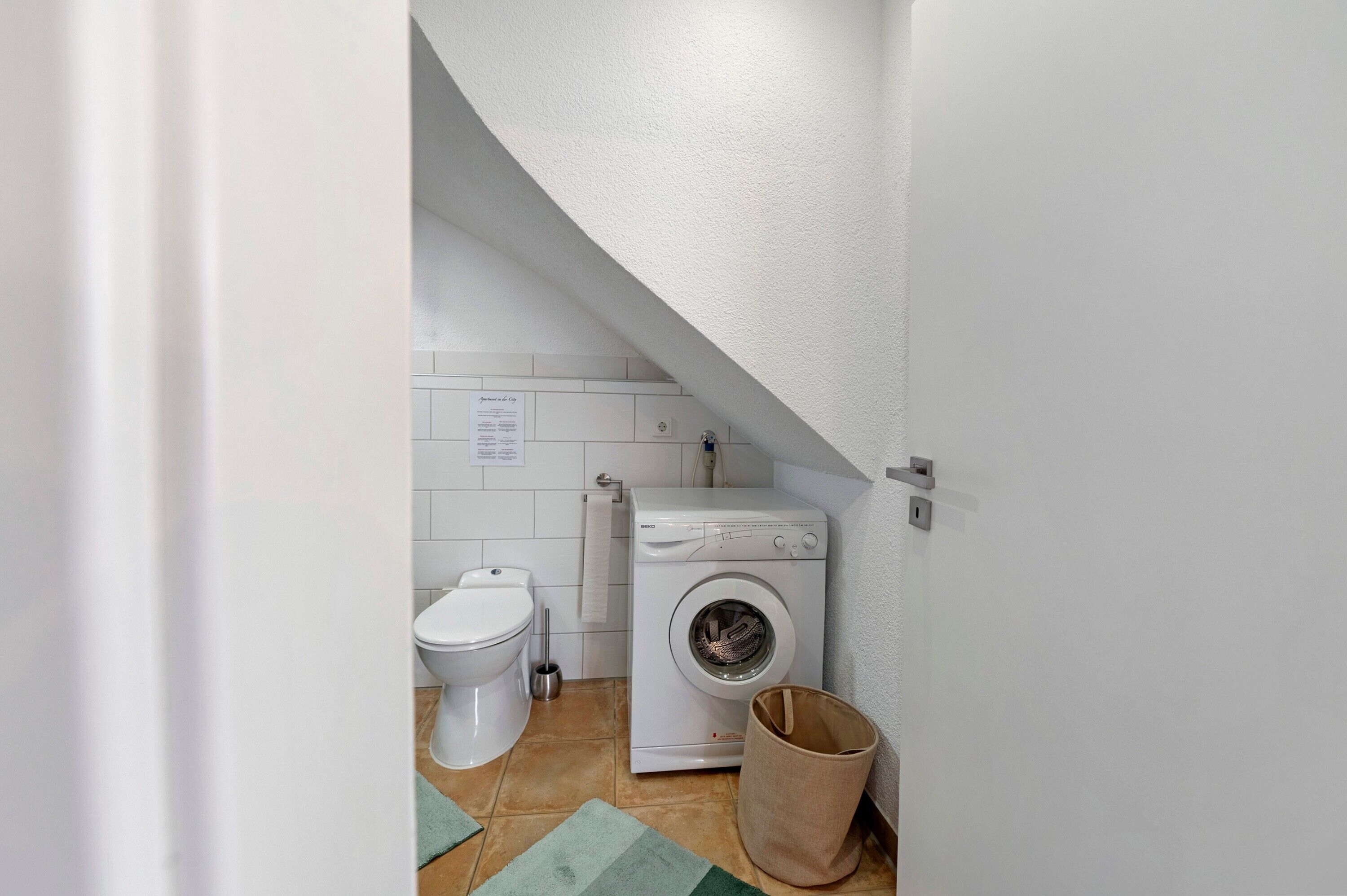 A small laundry room with a washing machine, a toilet, and a trash can.