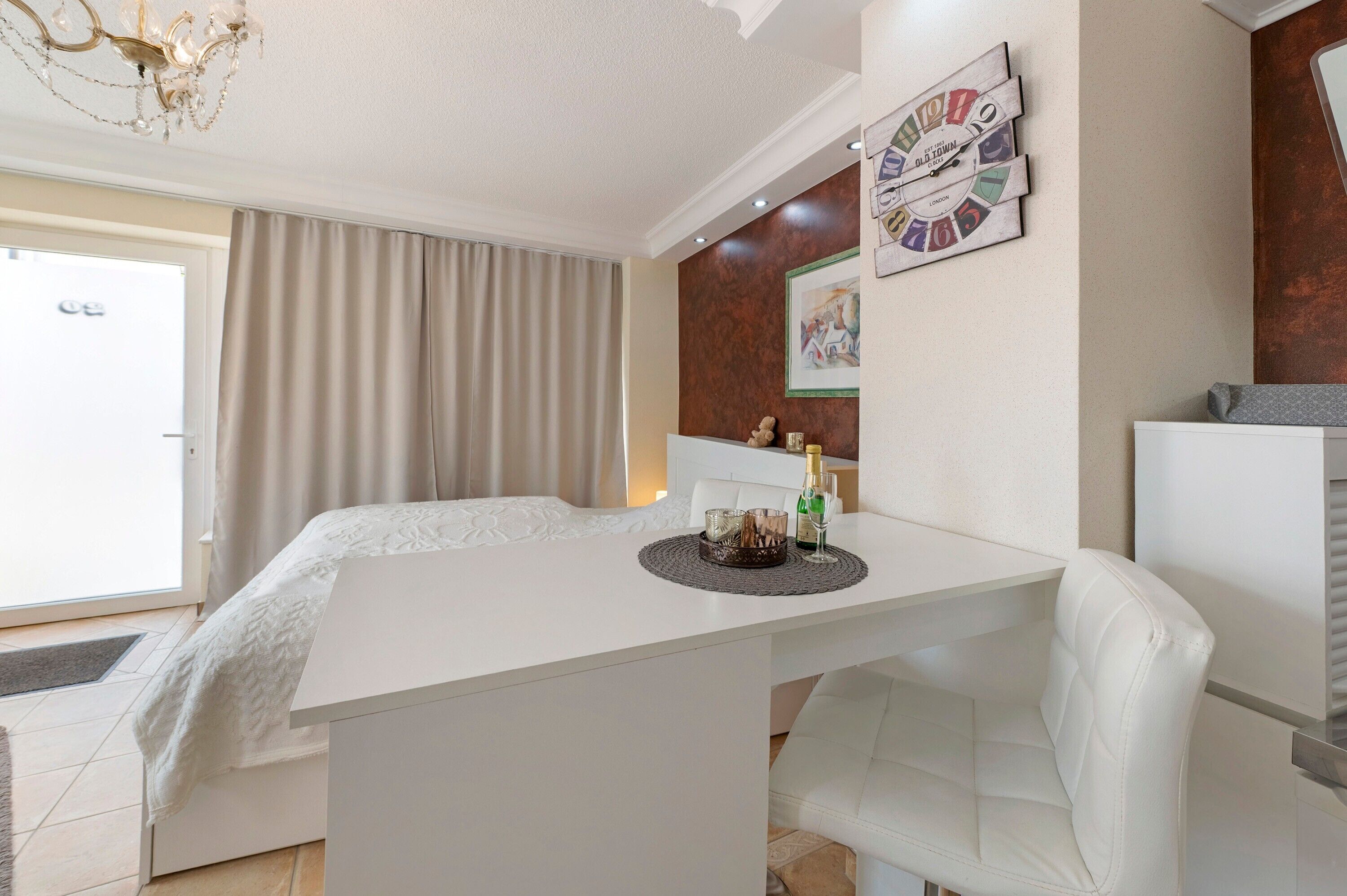 A room with a white bed, a dining table with chairs, a chandelier, a clock, and a window with curtains.