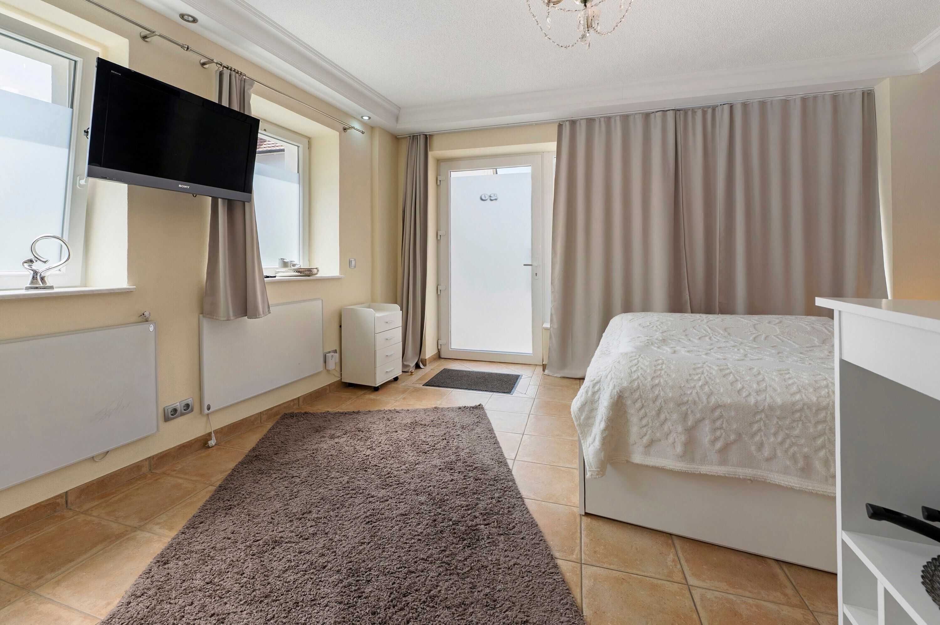 A bedroom with a bed, a TV mounted on the wall, a radiator, and a door leading to a balcony.