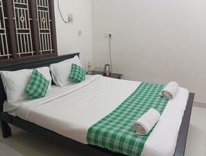 Una cama bien hecha con ropa de cama blanca y verde, una almohada y una toalla enrollada. Se ve una mesita de noche con una tetera y una botella.