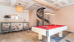 Une table de billard recouverte d’un drap de feutre rouge, un meuble ancien orné de motifs floraux, un escalier en colimaçon et un luminaire suspendu.