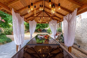 A covered outdoor dining area with a glass table, wooden ceiling, and hanging light fixtures. - Apartment 'Oiseaux' with Shared Pool, Wi-Fi and Air Conditioning - Villeneuve-lès-Montréal (Villeneuve-lès-Montréal)