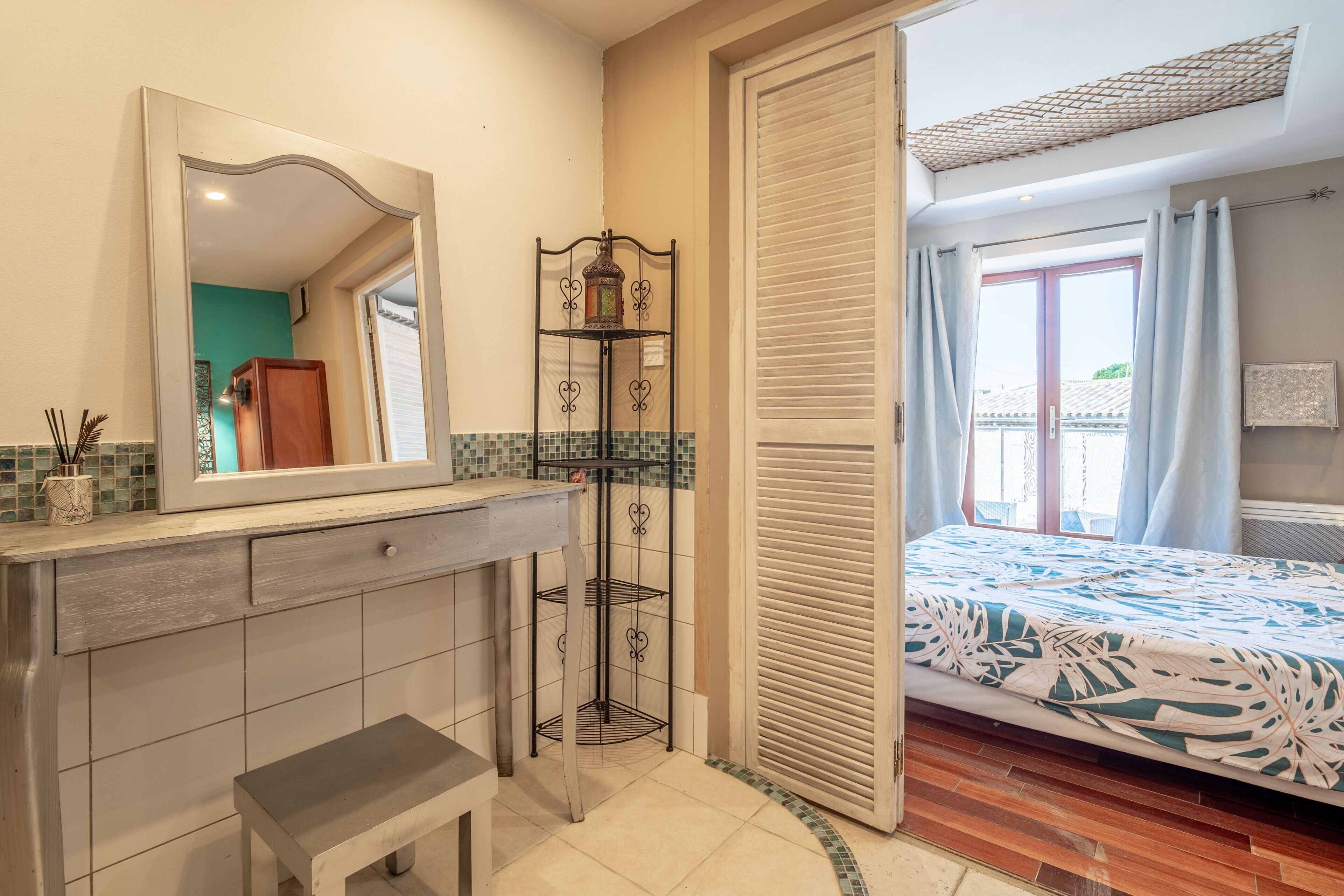Une salle de bain avec un miroir, un banc, une étagère avec une lanterne et un lit recouvert d’une couvre-lit à motifs.