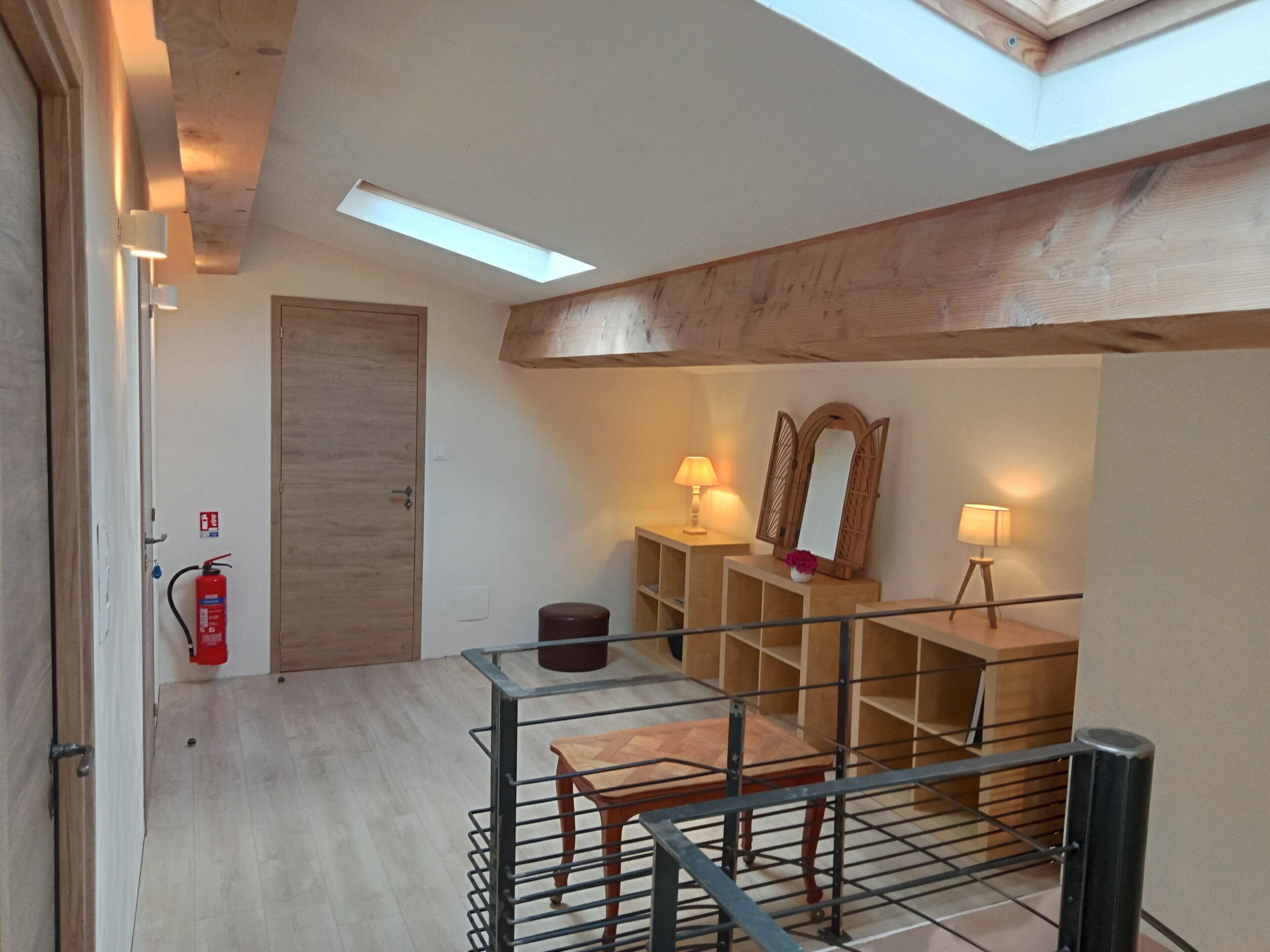 Une pièce de style « loft » avec une lucarne, des poutres en bois, un extincteur, un miroir et une petite table avec une lampe.