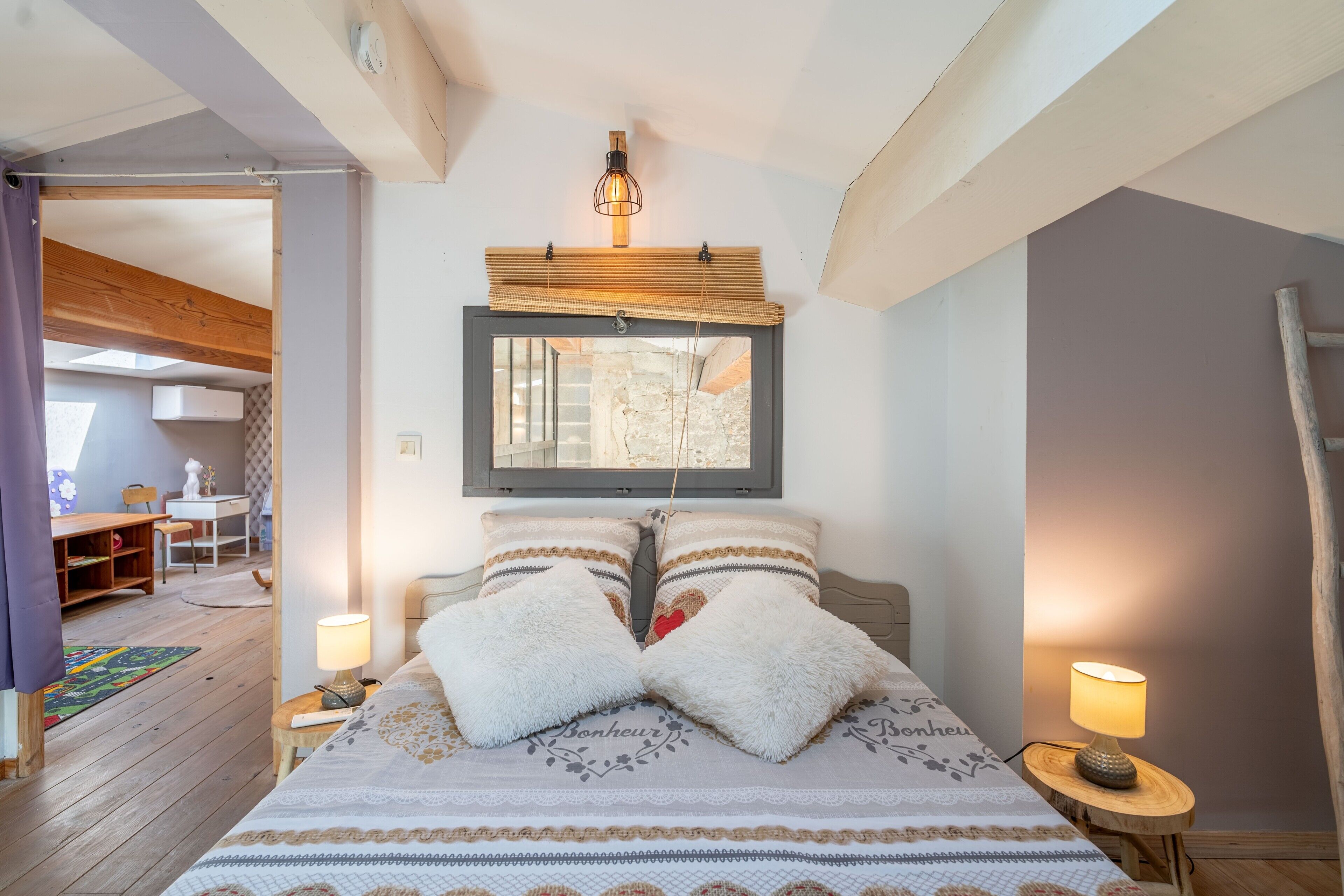 Une chambre à coucher comprenant un lit, des lampes de chevet, une tête de lit en bois, une fenêtre avec des stores et une vue sur un espace extérieur.
