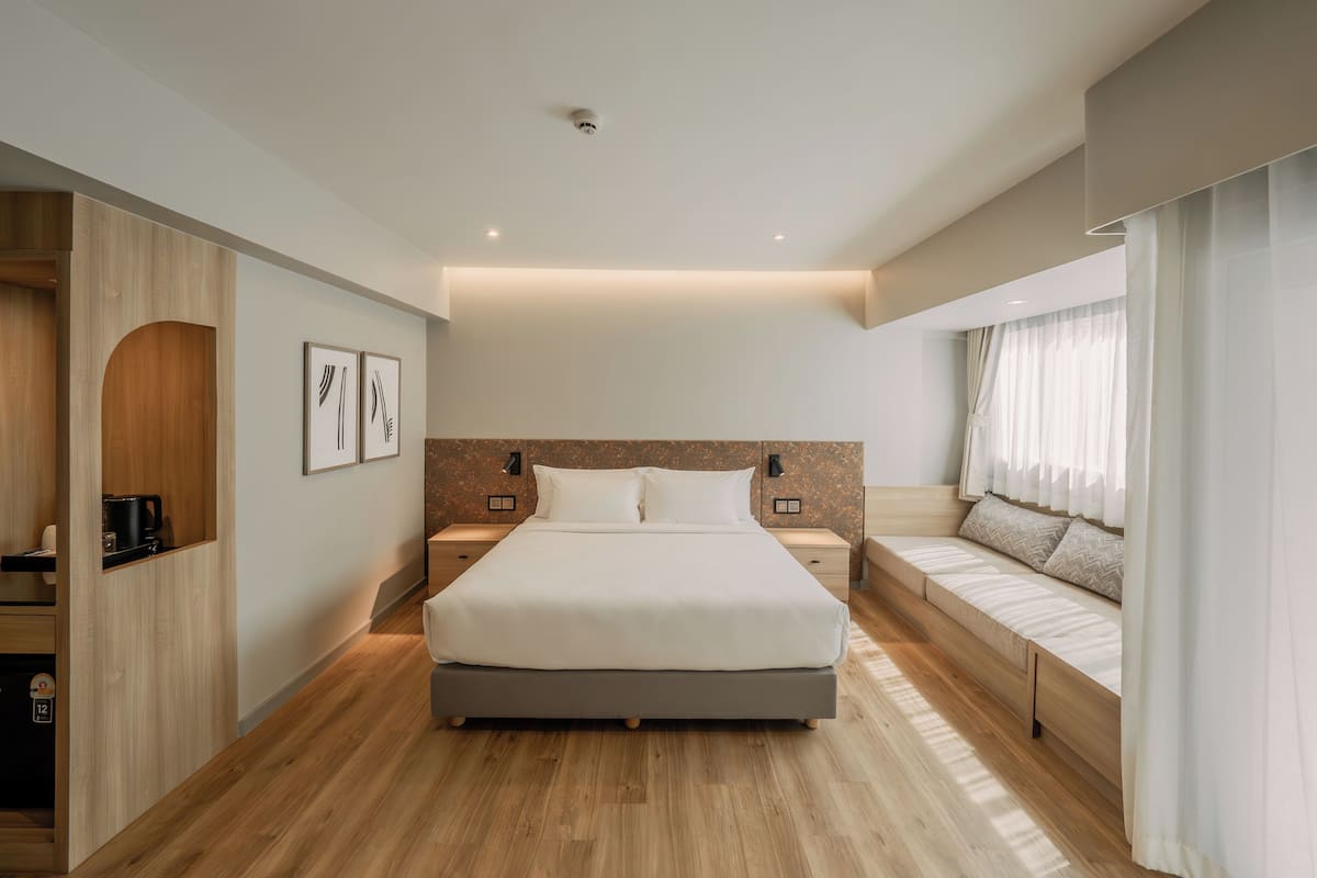 Una habitación de hotel moderna con una cama grande, mesitas de noche, un banco empotrado y vistas a la ciudad.