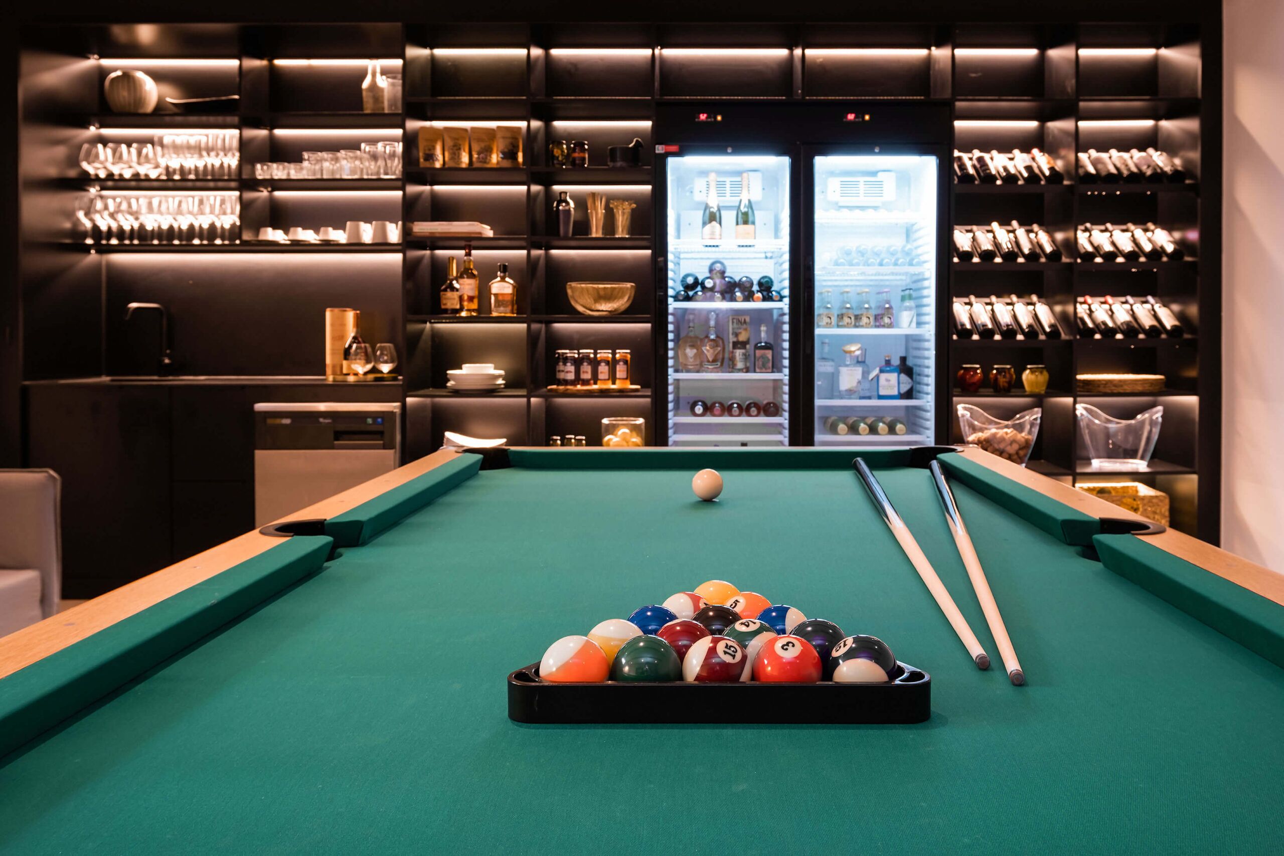 A pool table with balls and cues, surrounded by shelves of bottles and glasses.