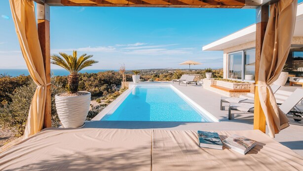 Une vue sur la piscine, avec un ciel bleu dégagé, une grande plante en pot et un livre posé sur une chaise longue.