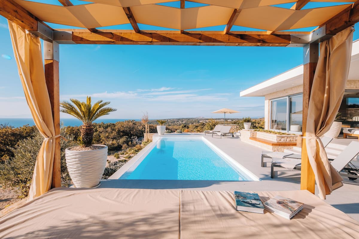 A poolside view with a clear blue sky, a large potted plant, and a book on a lounge chair.