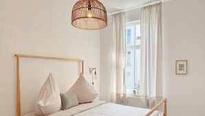 Ett minimalistiskt sovrum med en sängram i trä, vita sängkläder, ett nattduksbord, en hängande lampa och ett fönster med tunna, genomskinliga gardiner.