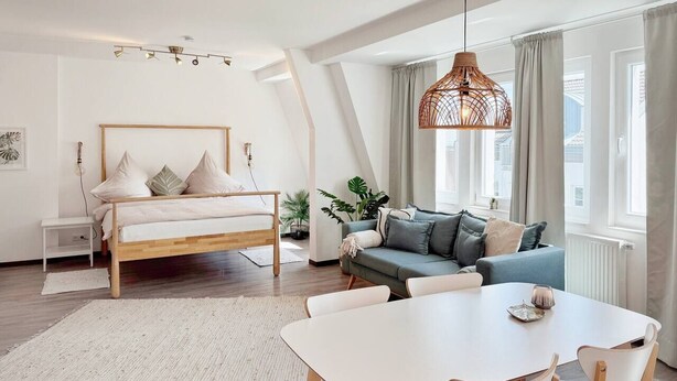 A modern bedroom with a wooden bed, a gray sofa, a dining table with chairs, and a hanging light fixture.