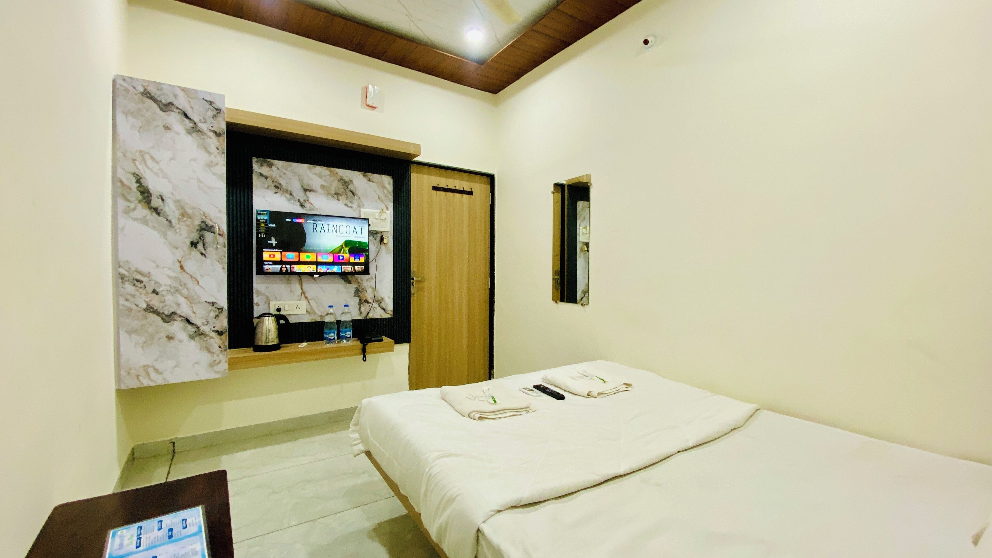 A hotel room with a bed, a TV mounted on a marble wall, a wooden door, and a small table with a kettle and bottles.