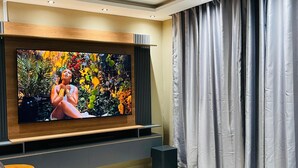 A modern living room with a large flat-screen TV mounted on a wooden panel, grey curtains, and a brown leather sofa.