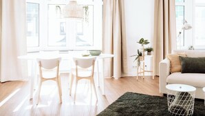 A bright, modern living room with a white sofa, a round dining table with two chairs, a hanging light fixture, and a small side table with plants.
