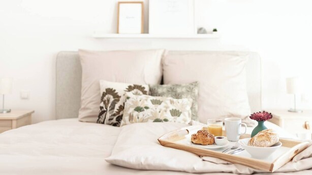 A neatly made bed with a tray of breakfast items, including croissants, coffee, and juice, in a bright, minimalistic room with a white color scheme.