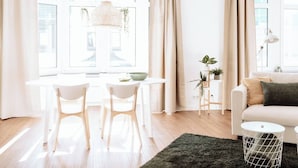 A bright, modern living room with a white sofa, a round dining table with two chairs, a hanging light fixture, and a small side table with plants.