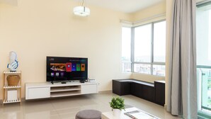 A modern living room with a flat-screen TV, a white coffee table, a grey ottoman, and a large window with a view of the city.