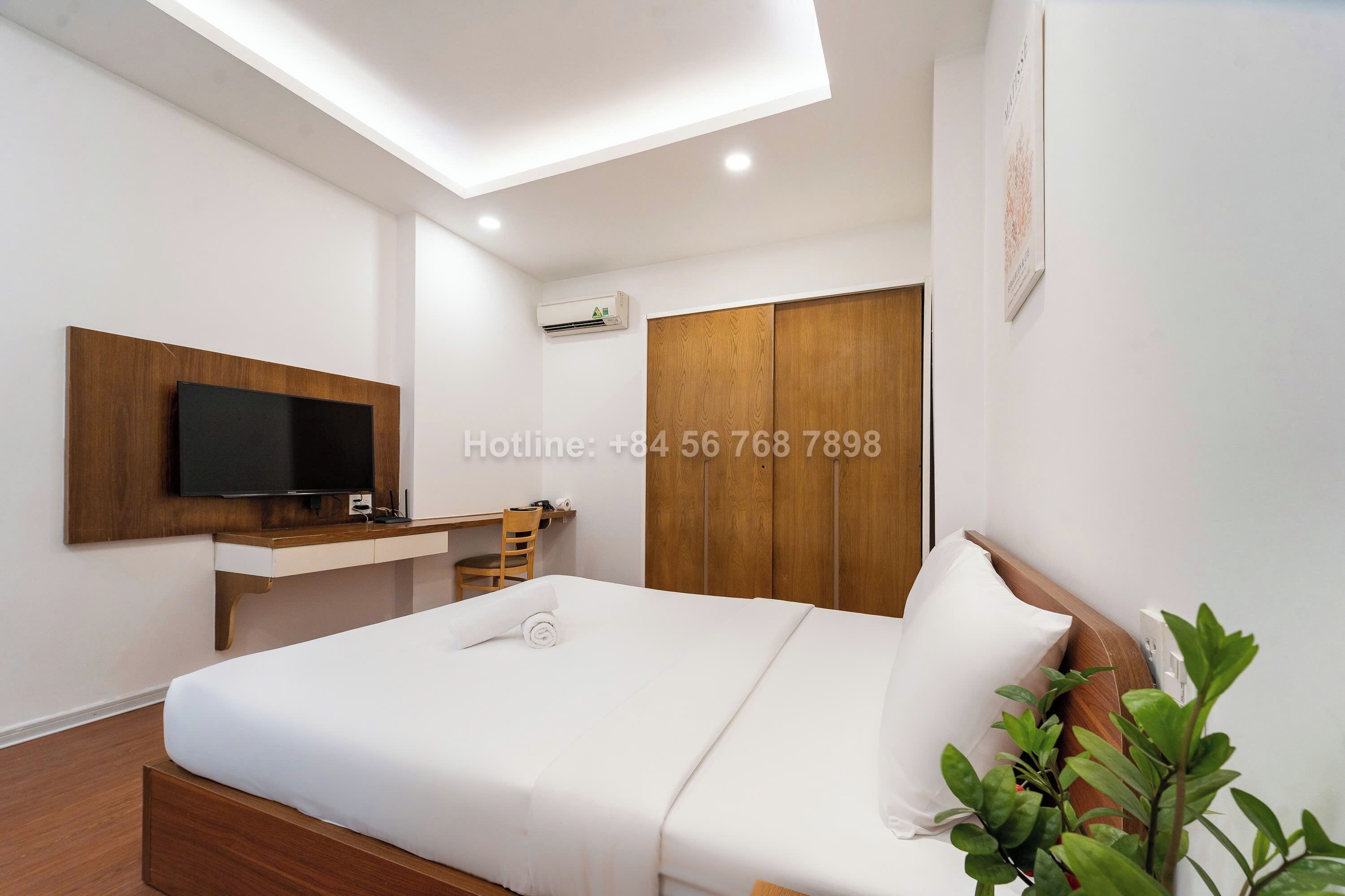 A modern hotel room with a bed, a TV mounted on a wooden panel, a desk, and a wardrobe.