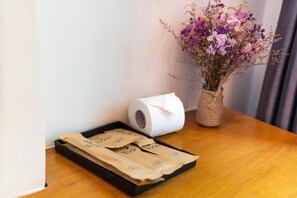 Une table en bois, un rouleau de papier toilette, un vase avec des fleurs séchées et un plateau avec des sacs en papier.