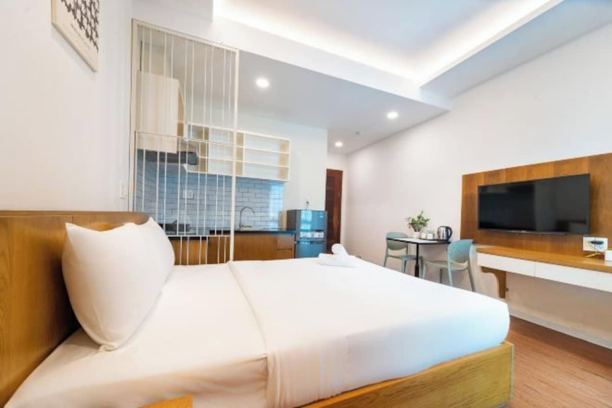 A modern hotel room with a bed, a desk, a TV, and a kitchenette.
