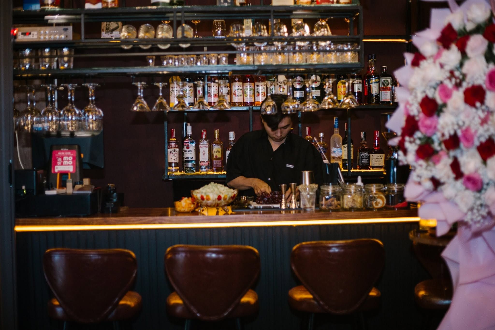 A bar with a bartender preparing drinks, shelves of liquor bottles, and bar stools.
