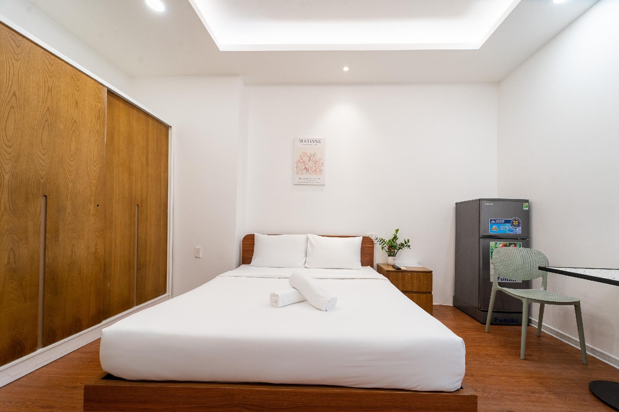 A hotel room with a bed, a desk, a chair, a refrigerator, and a wall-mounted artwork.