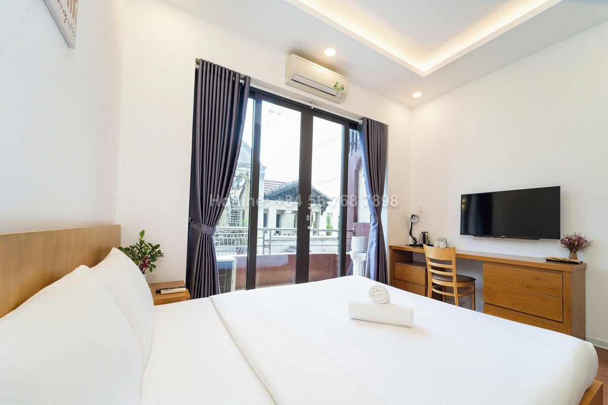 A hotel room with a bed, a desk, a chair, a TV, and a balcony with curtains.