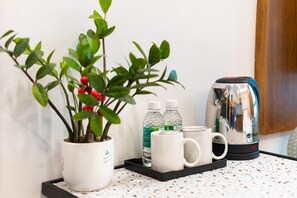 Un petit coin cuisine équipé d’une machine à café, de deux bouteilles d’eau, de deux tasses blanches et d’une plante en pot aux feuilles vertes et aux baies rouges.