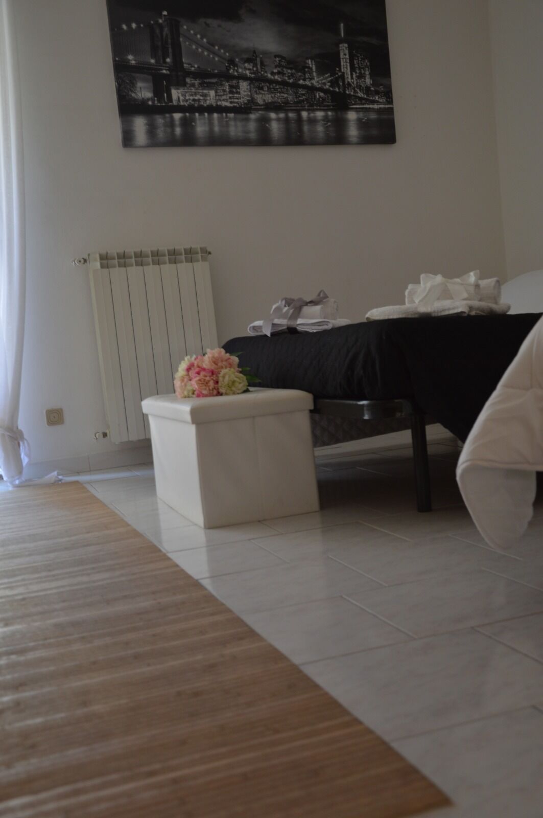 A hotel room with a bed, a radiator, a small table with a bouquet of flowers, and a painting of a cityscape on the wall.