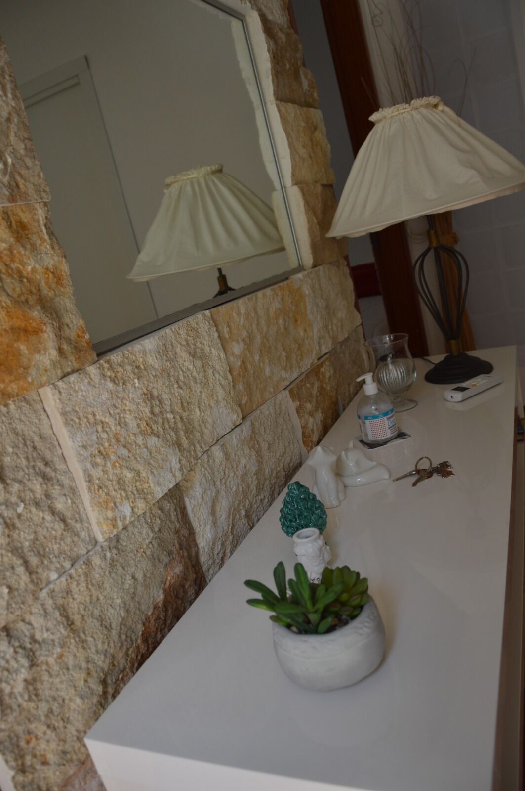 Un baño con pared de piedra, un espejo, un lavamanos blanco, una planta en maceta, un dispensador de jabón y una lámpara.