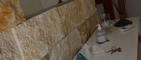 A bathroom with a stone wall, a mirror, a white sink, a potted plant, a soap dispenser, and a lamp.