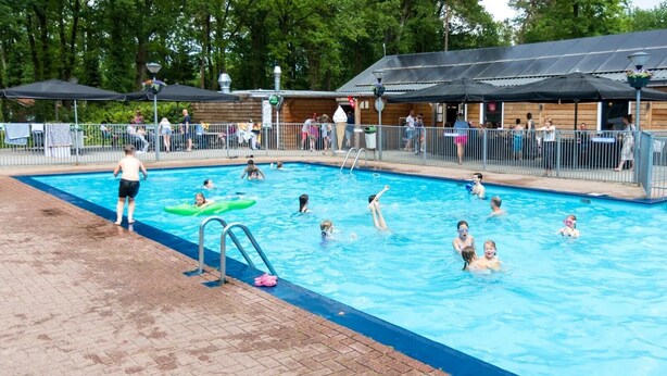 A swimming pool with people in the water, a lifeguard, and a covered area with a building in the background. - The Molshoop - Detached nature cottage in the middle of the Forest (IJhorst)
