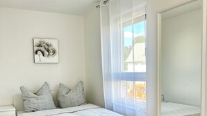 A bedroom with a bed, a bedside table, a nightstand, a window with sheer curtains, a mirror, and a wall-mounted picture. - Sun oasis in Sigmaringen (Sigmaringen)