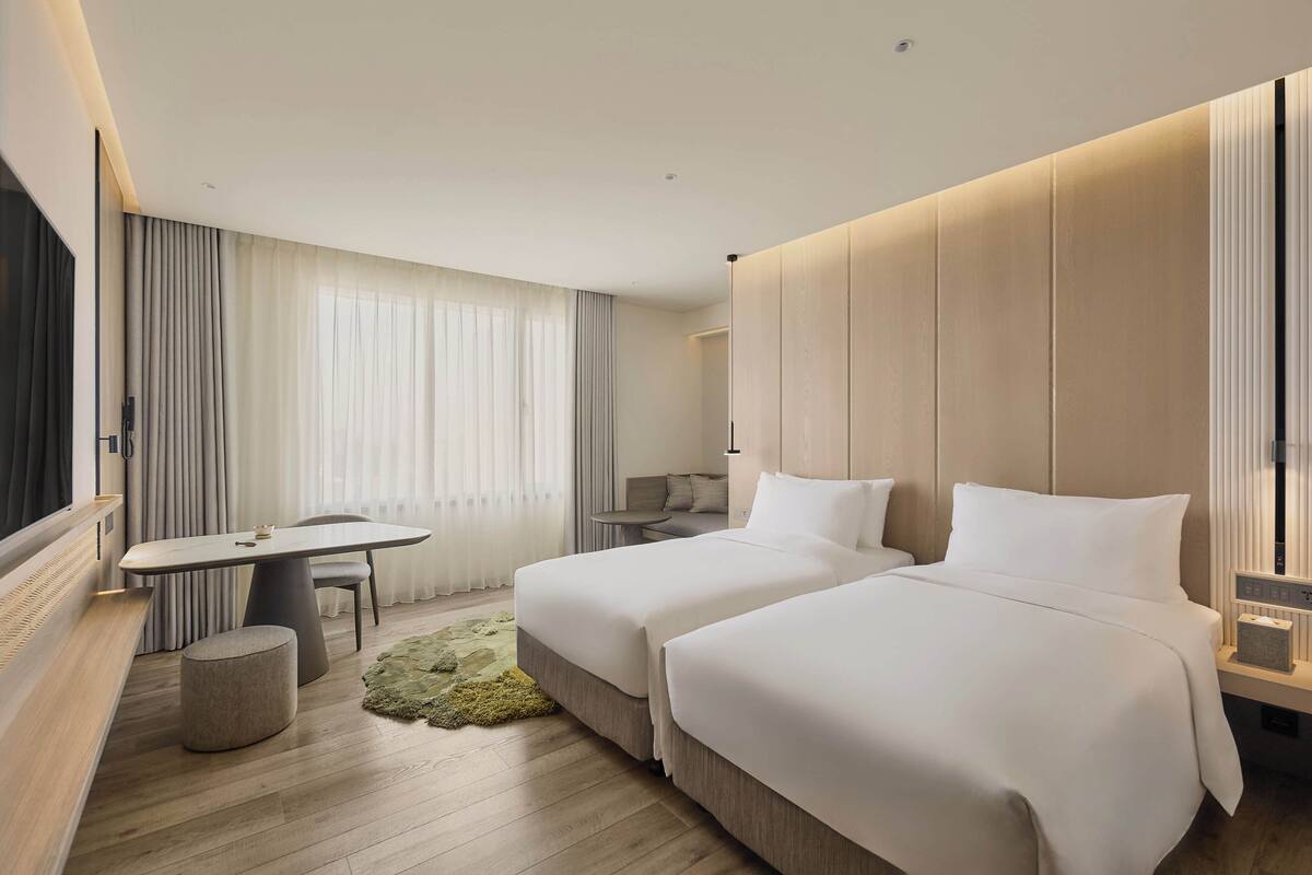 A hotel room with two beds, a dining table, a small round table, a bench, and a large window with curtains.