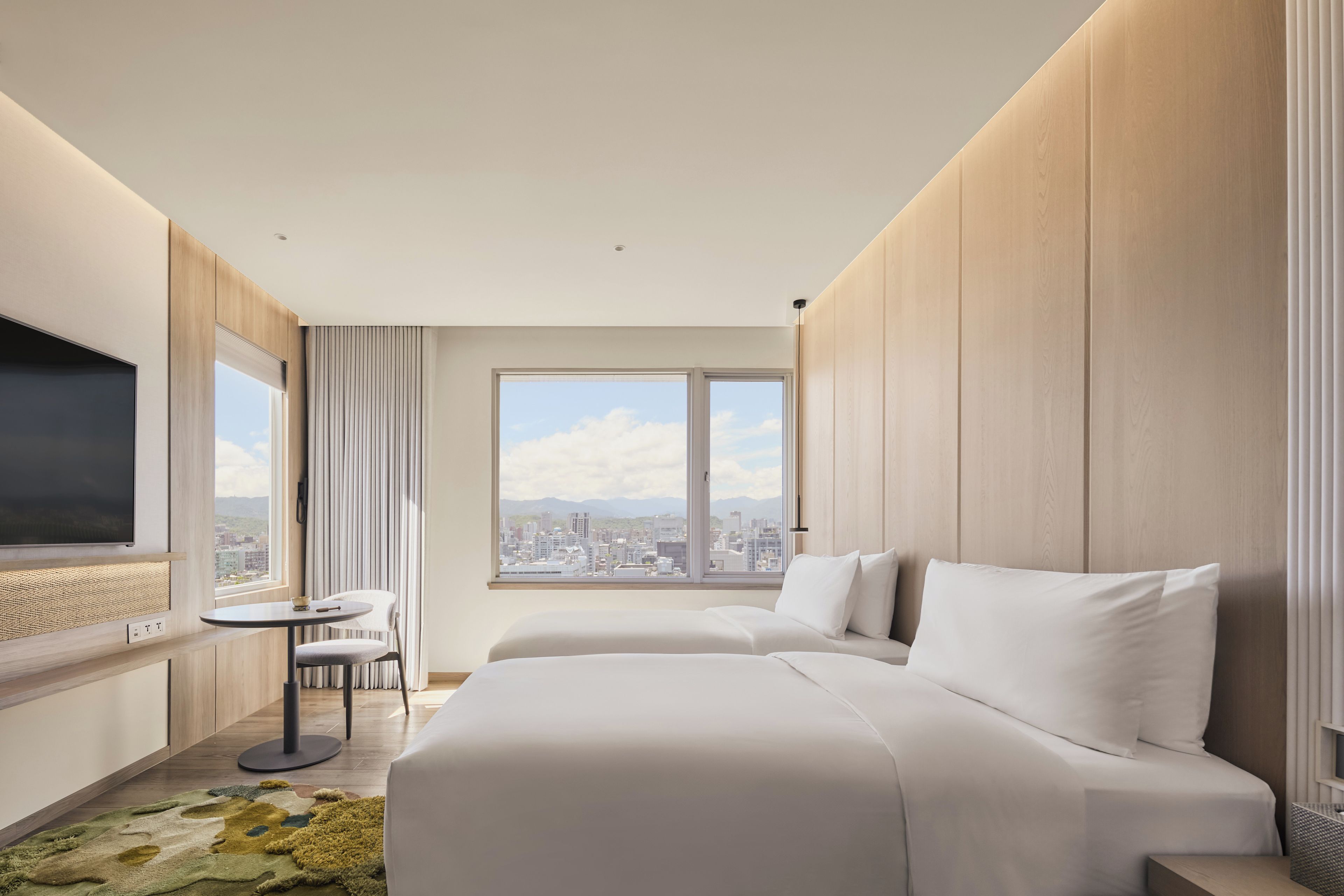 A hotel room with a large bed, a small table, a chair, a TV, and a window with a city view.