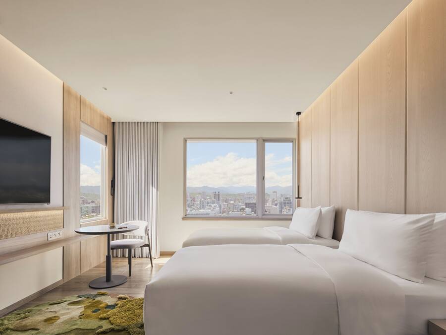 A hotel room with a large bed, a small table, a chair, a TV, and a window with a city view.