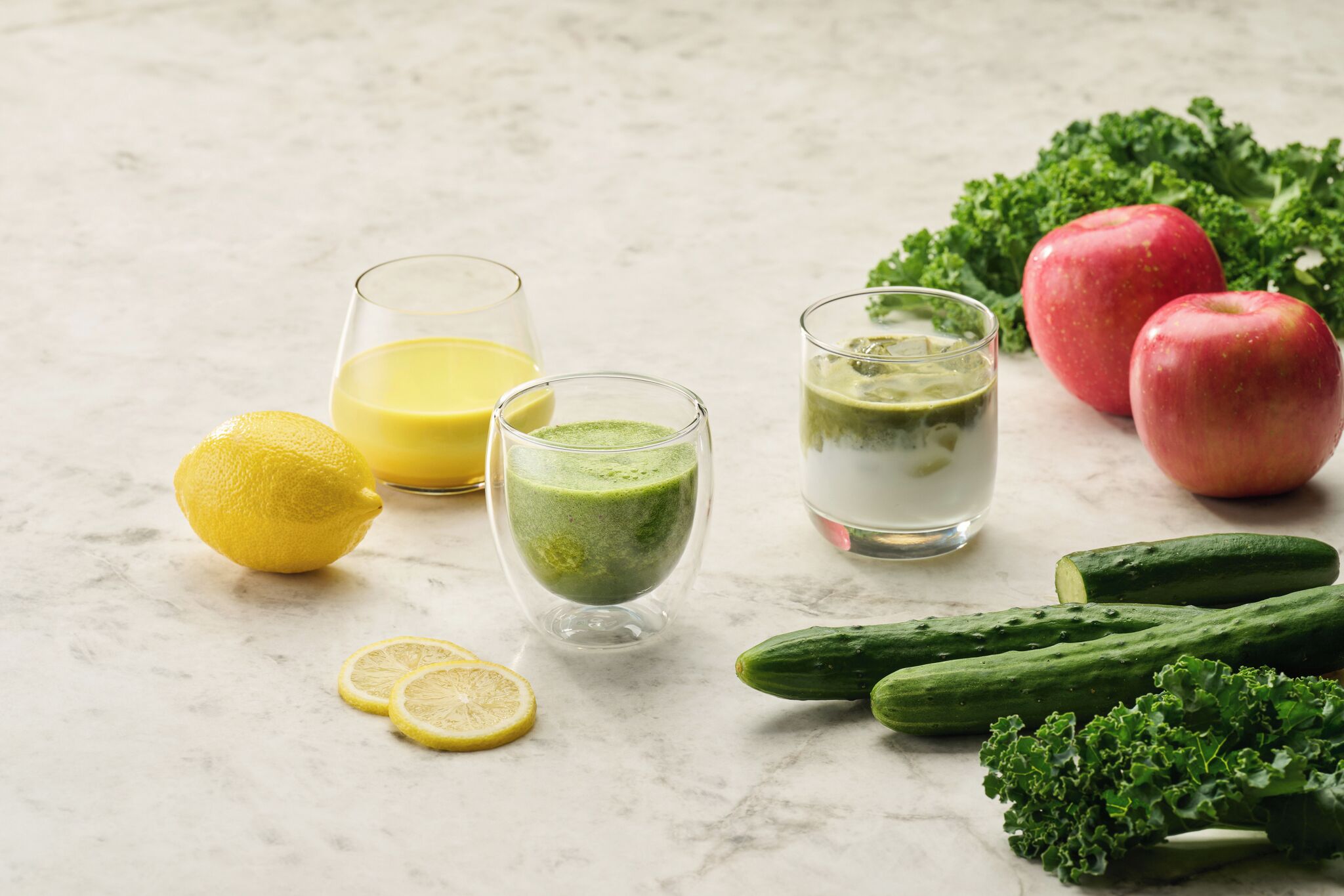 Una mesa con tres vasos de jugo verde, un limón y verduras como pepinos y col rizada.
